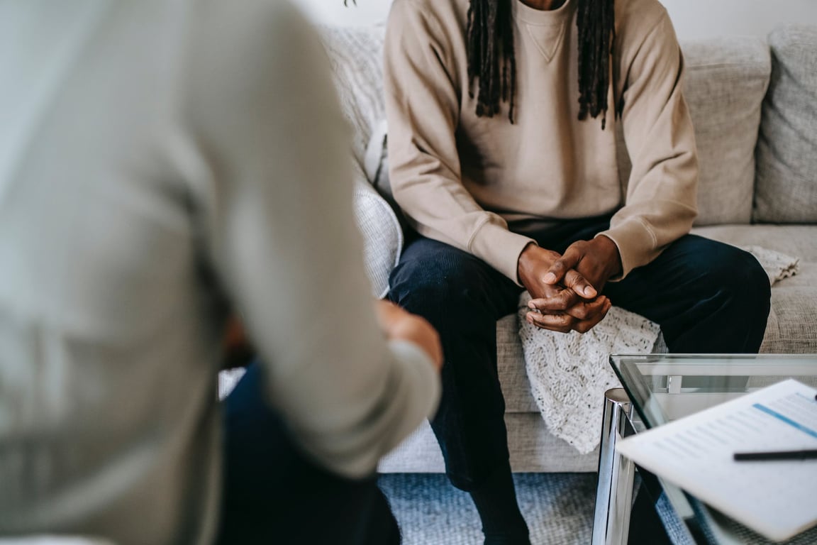 Patient afro-américain méconnaissable, les mains jointes, assis sur un canapé près d'un thérapeute anonyme et flou lors d'une visite dans un cabinet de psychothérapie.